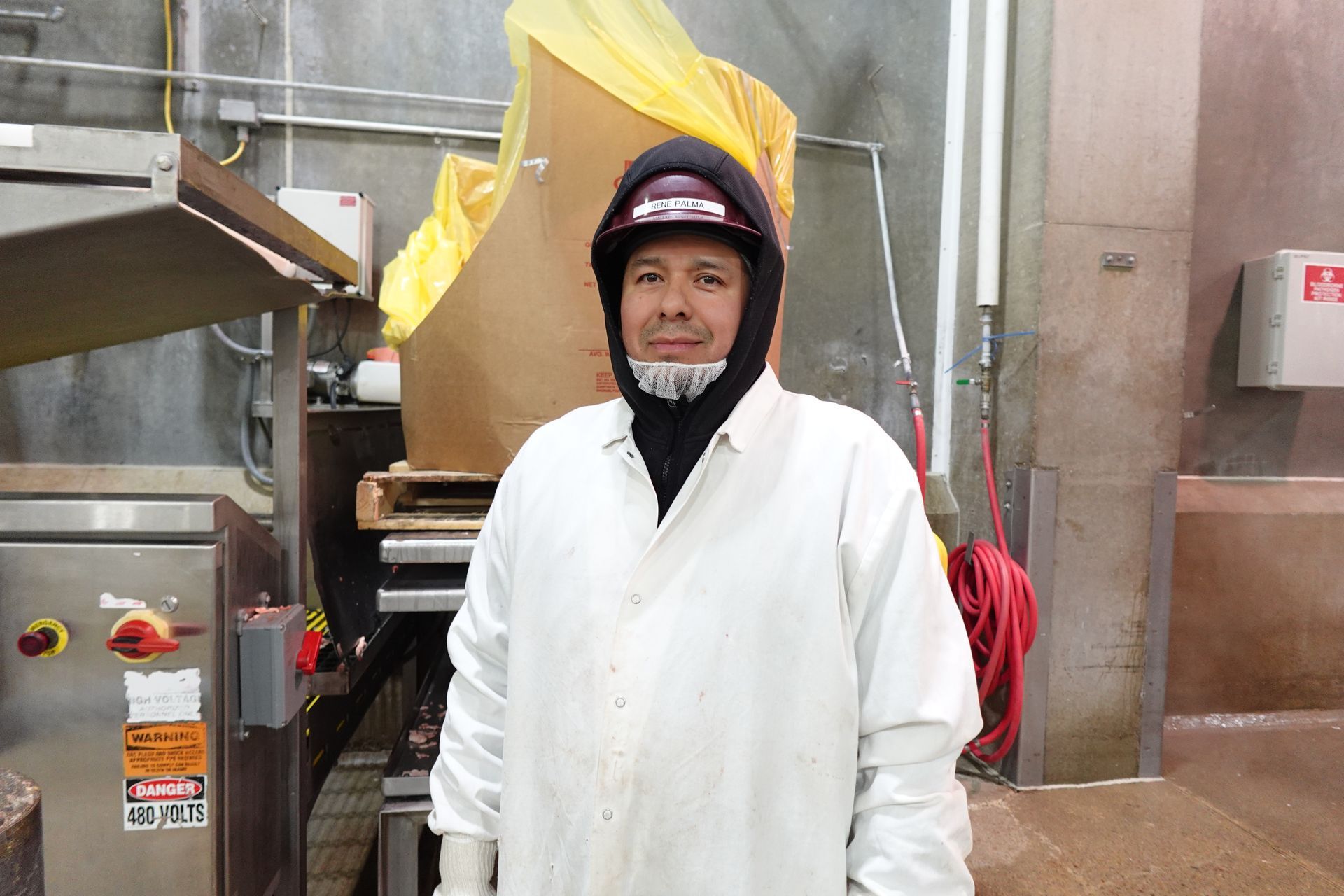 Person in a white coat and hard hat standing in an industrial kitchen, smiling at the camera.