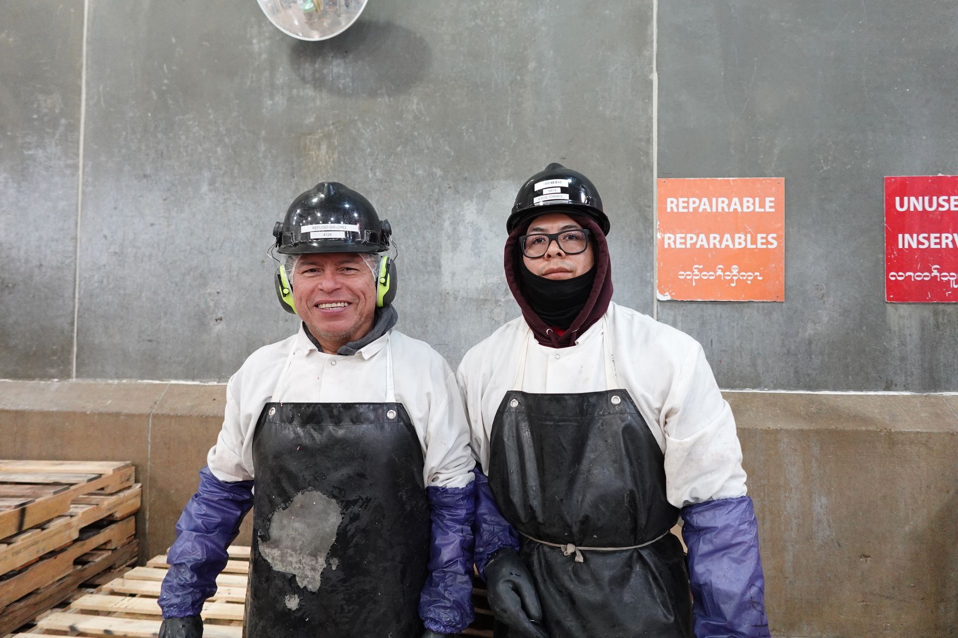 Two workers in white protective suits and black aprons standing indoors, smiling in front of a concrete wall.