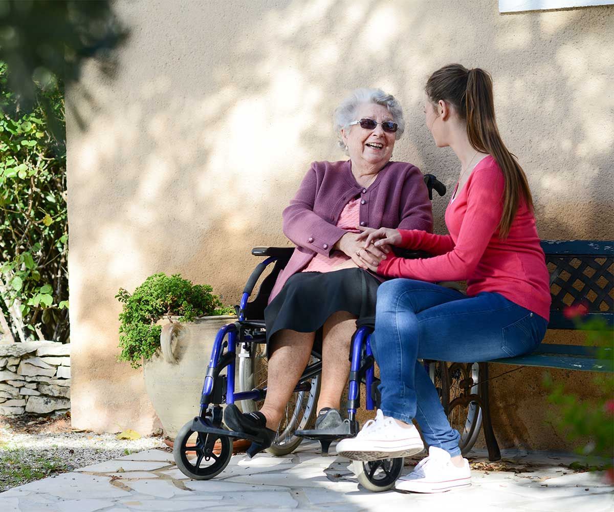 An older person in a wheelchair smiles at a younger person sitting on a bench, holding hands. Outdoors.