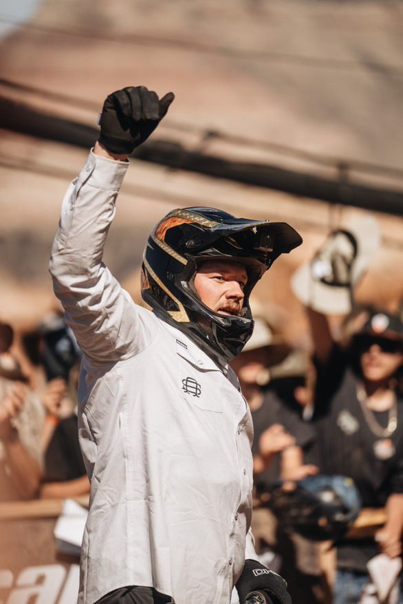 Person in helmet raises arm in air, celebrating. Crowd blurred in background, sunny outdoor setting.