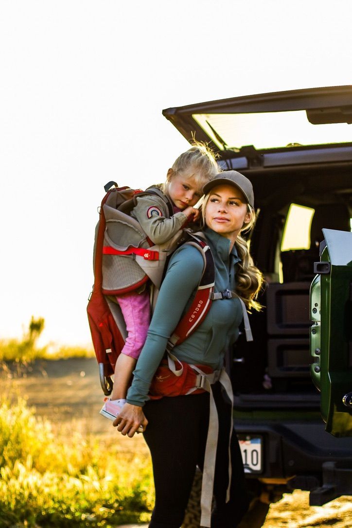 Woman with a child in a carrier, hiking near a vehicle. Warm sunlight, blonde hair.