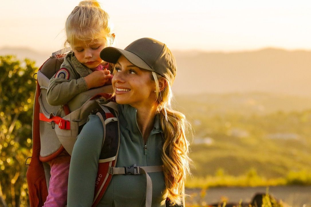Woman hiking with a child in a carrier, both looking towards sunlit hills.