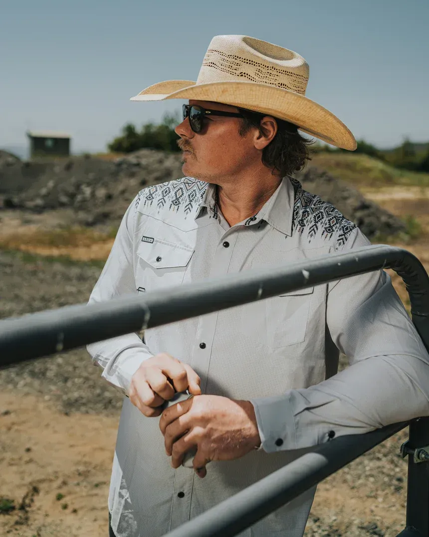 Man in cowboy hat and sunglasses leans on a metal gate, looking off to the side.