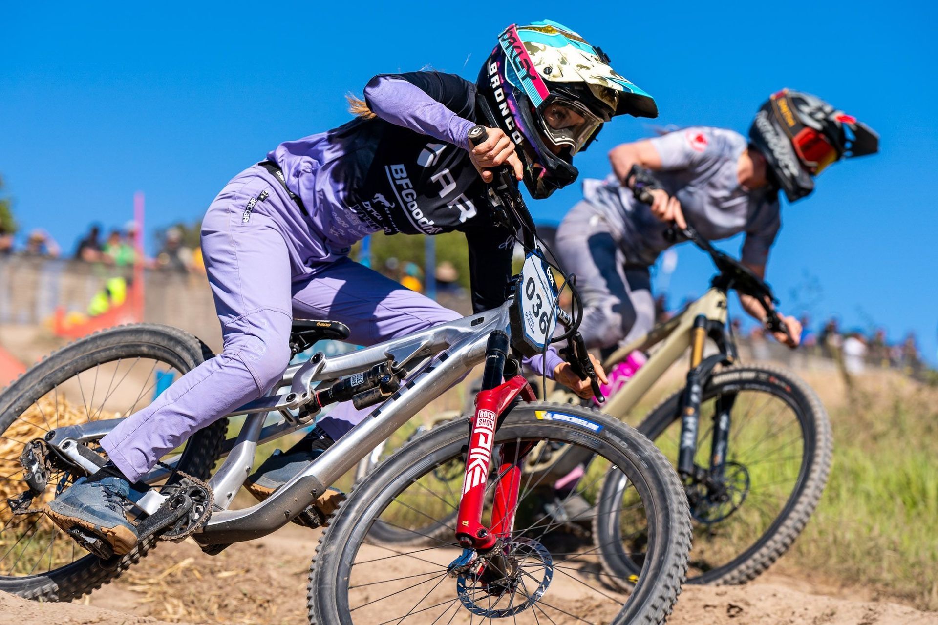 Two women on mountain bikes racing downhill on a dirt track, sunny day.