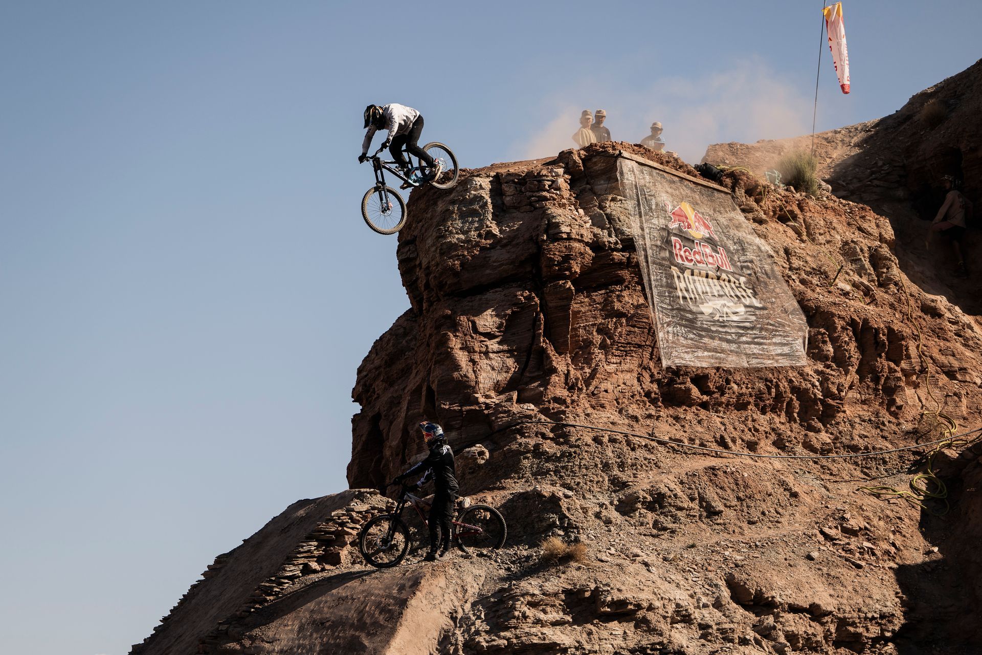 Mountain biker launching off a cliff, another biker below, Red Bull banner, desert setting.