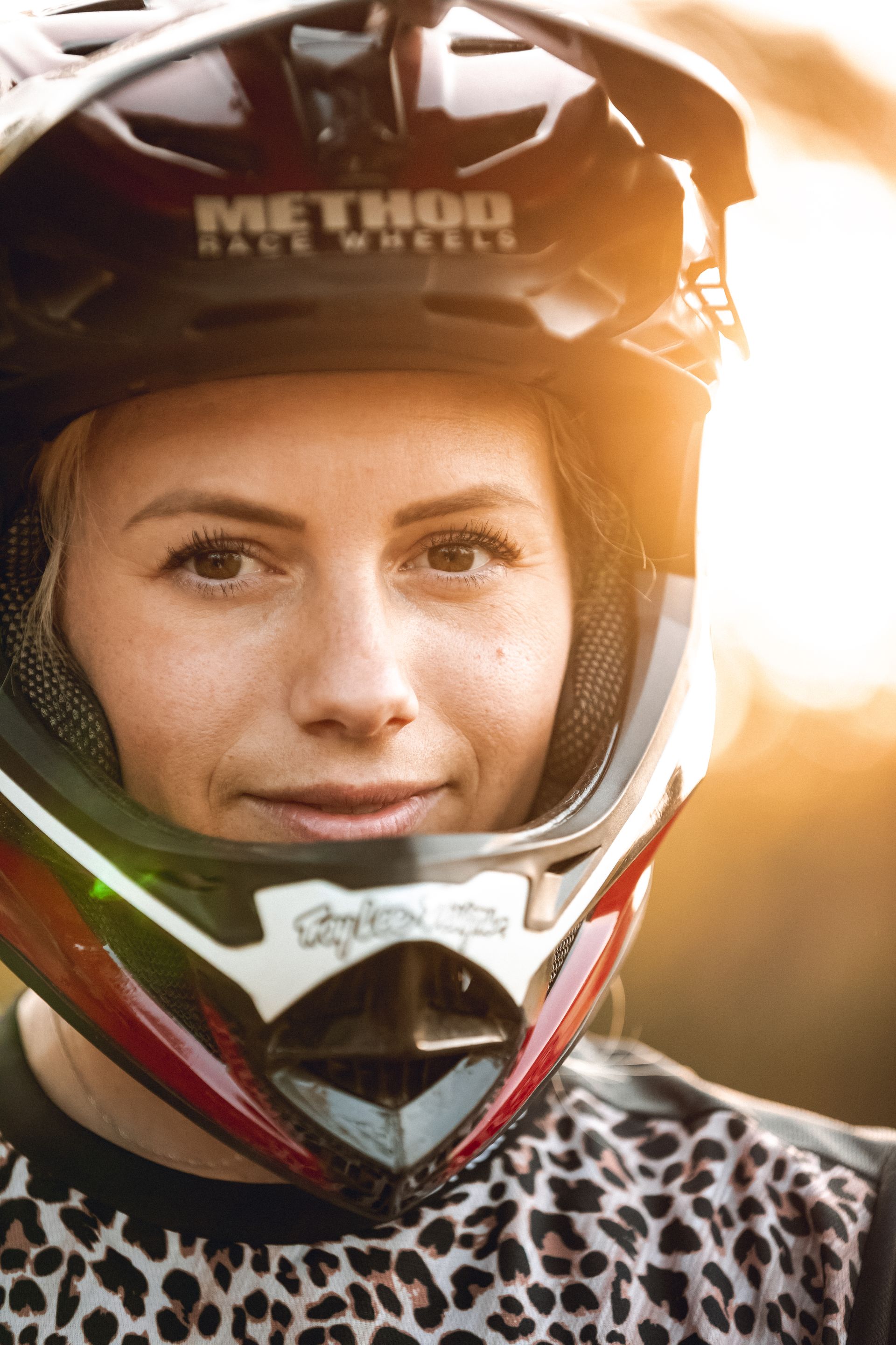 Woman wearing a motocross helmet, smiling, with sunlight in the background.