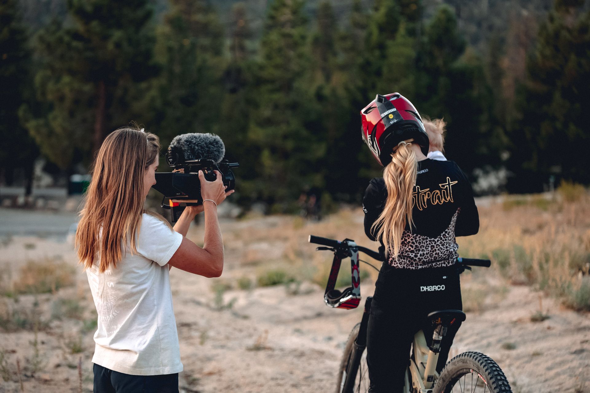 Woman filming a mountain biker with a helmet; setting is outdoors near trees.