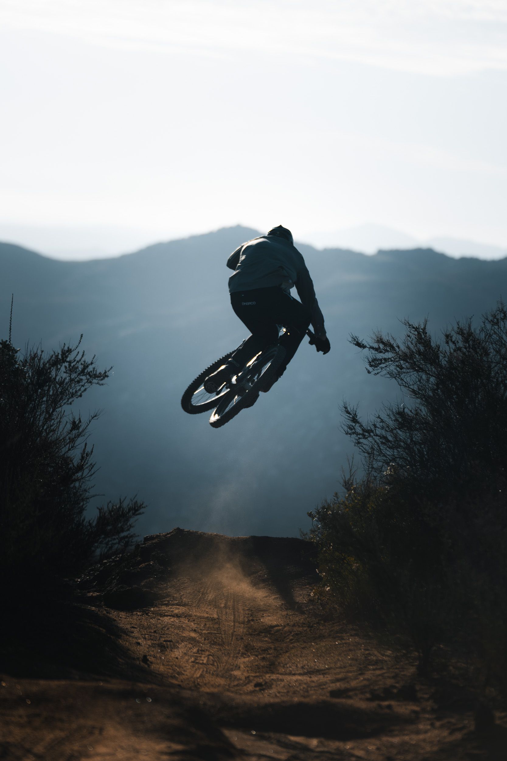 Mountain biker jumps over a dirt trail, silhouetted against a hazy mountain backdrop.