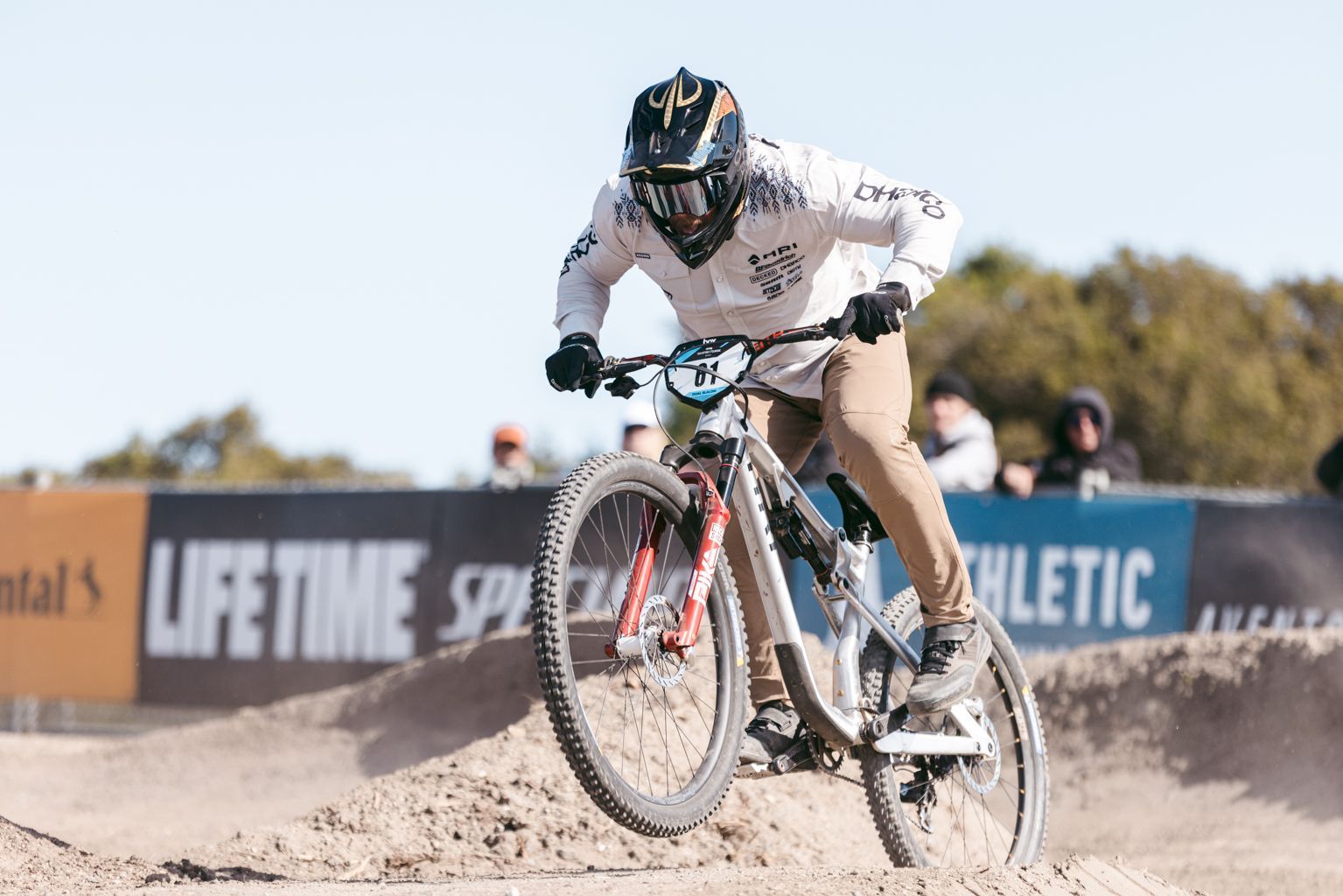 Mountain biker in mid-air, riding a dirt track. He wears a helmet, white top, brown pants, and rides a Specialized bike.
