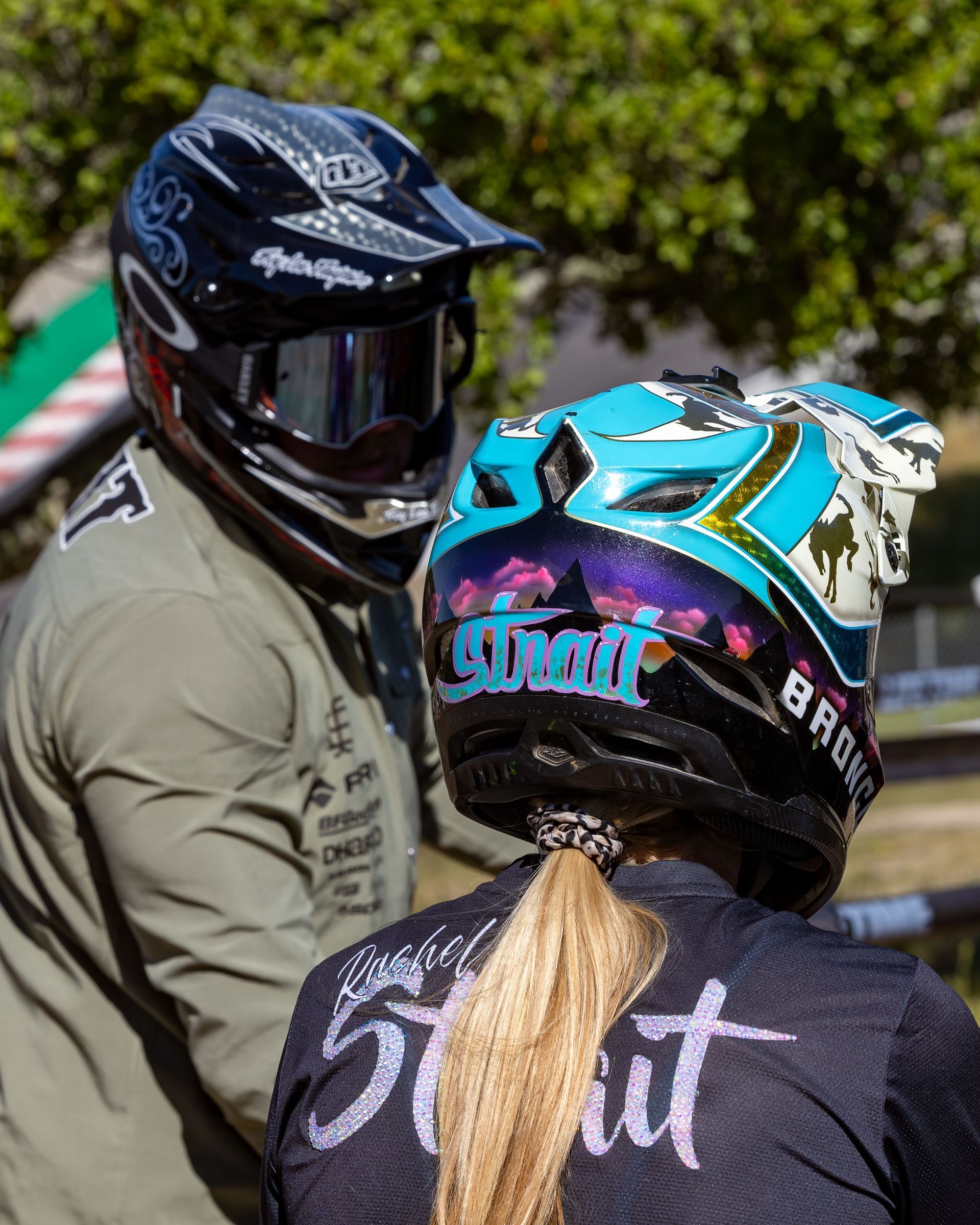 Person in helmet raises arm in air, celebrating. Crowd blurred in background, sunny outdoor setting.