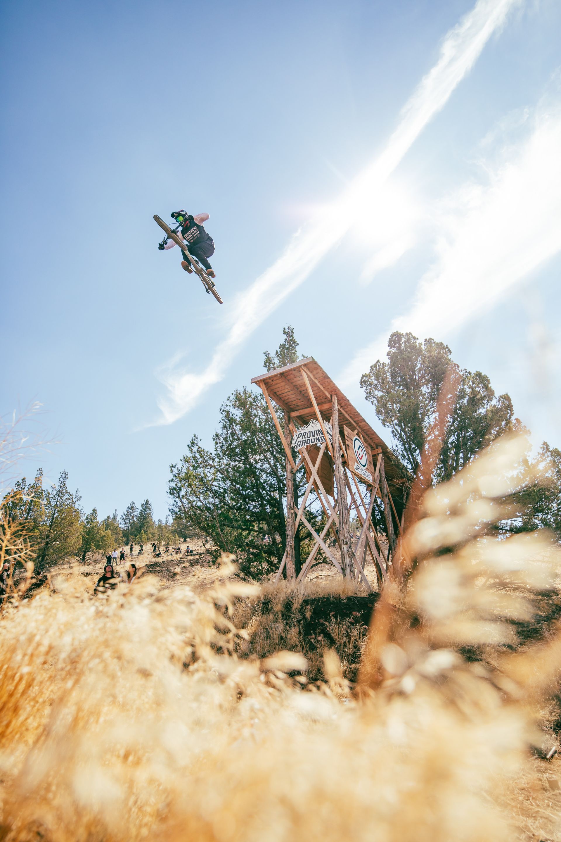 Snowboarder in mid-air over wooden ramp. Sunny day, blue sky, brown grass and trees.