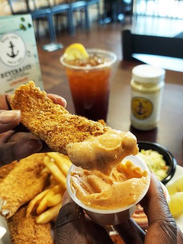 Person dips fried fish into sauce. Fries, coleslaw, iced tea, and jar of sauce on a table.