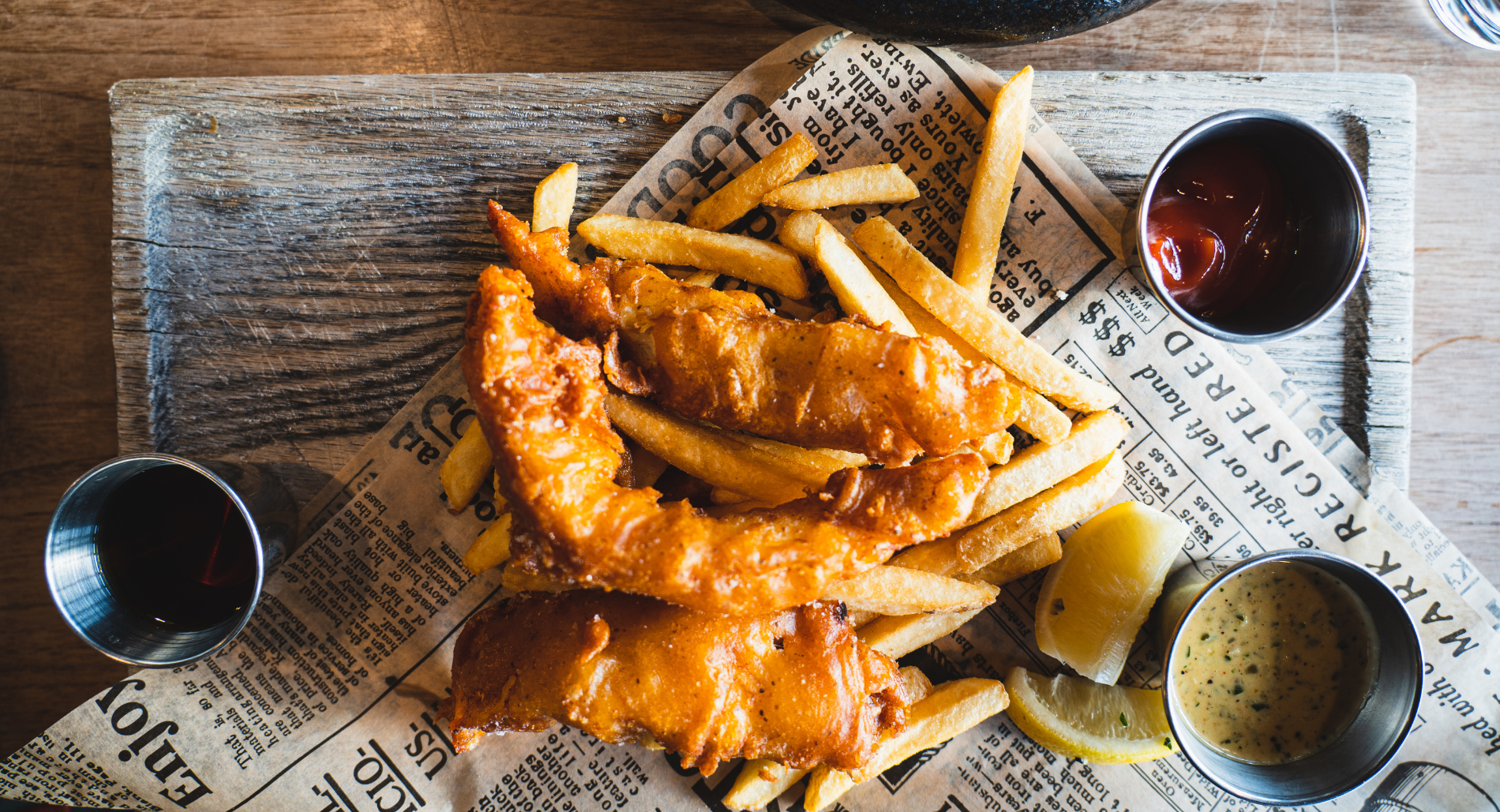 Fish and chips with dipping sauces on a wooden tray lined with newspaper.