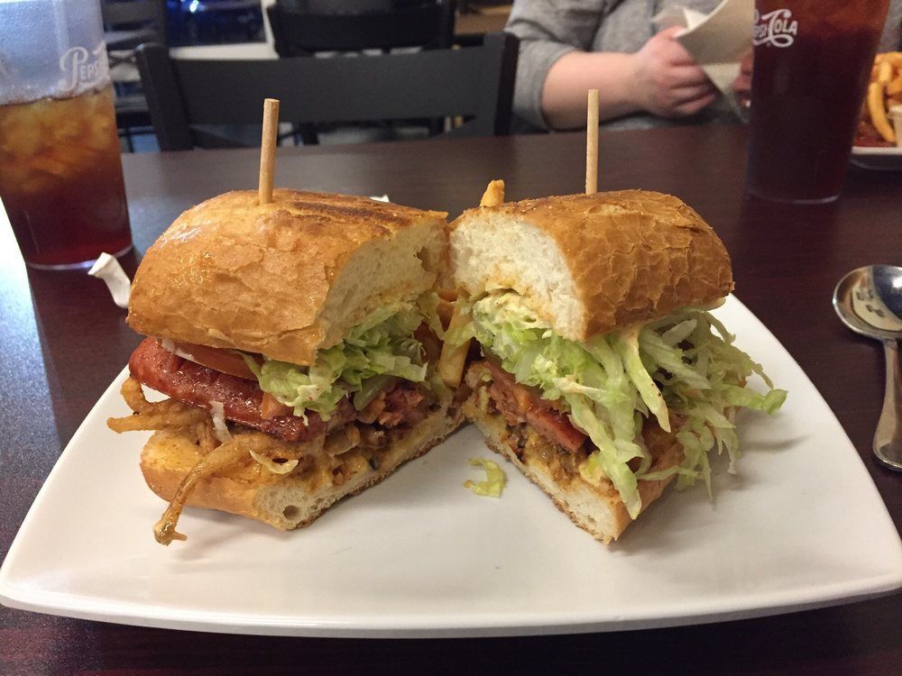 A halved, loaded sandwich on a white plate, with a drink and person in the background.