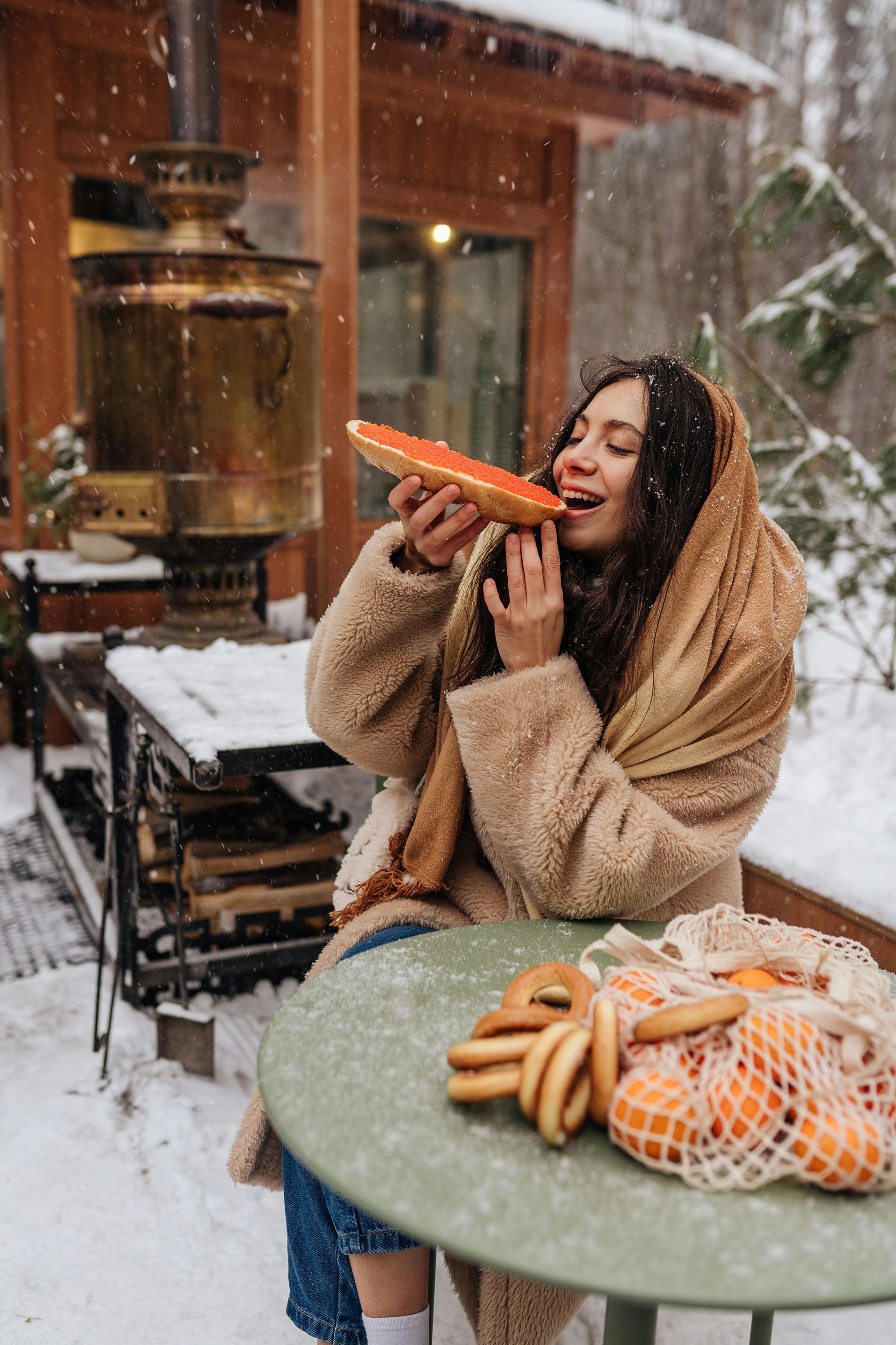 Woman enjoying food in snowy setting, wearing scarf and coat. Bagels and oranges on table, samovar in background.