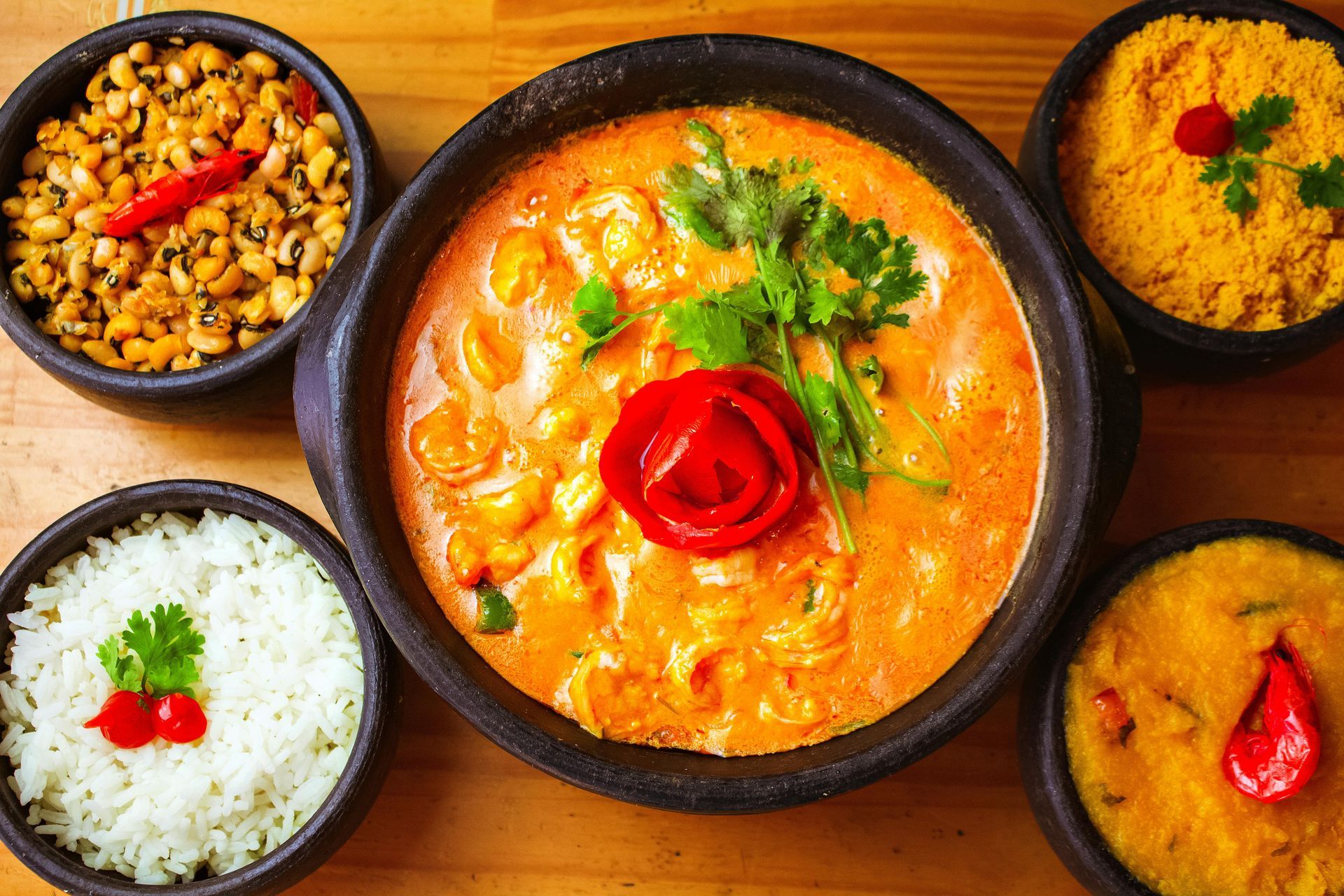 A bowl of creamy shrimp moqueca centered among side dishes of black-eyed peas, white rice, farofa, and a yellow puree.