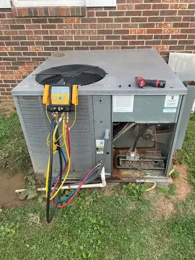 HVAC unit being serviced with gauges and hoses connected, outside on grass, brick wall in background.