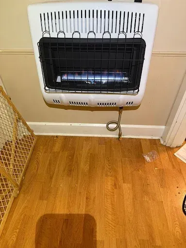 White wall-mounted gas heater with a blue flame. It sits above wooden flooring.