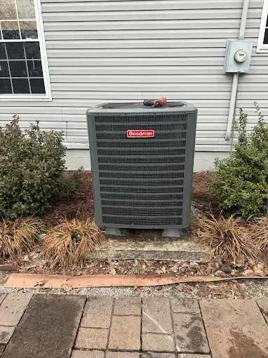 An outdoor Goodman AC unit on a concrete pad next to a building with bushes and a brick patio.