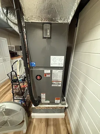 HVAC unit in a small room next to a white-paneled wall and a small cabinet. Fan in the foreground.