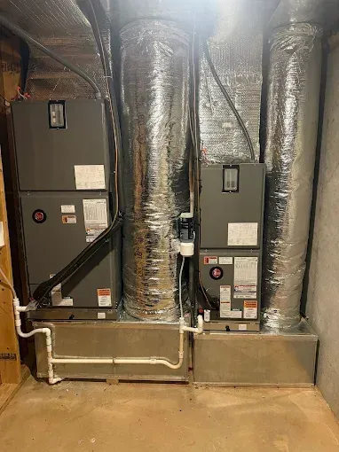 Two gray HVAC units with silver ductwork in a basement setting. White PVC pipes connect to the units.