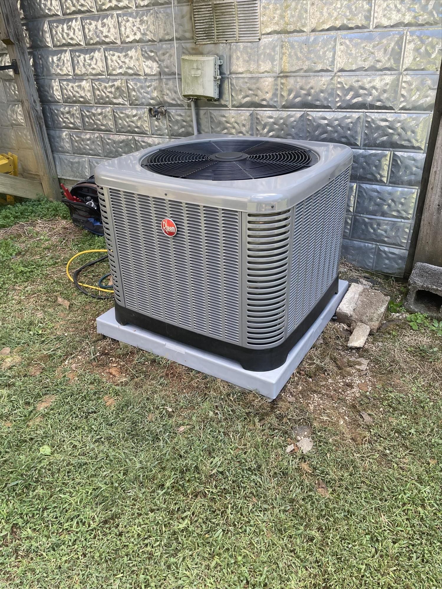 Air conditioning unit on a concrete pad in a grassy yard, against a brick wall.