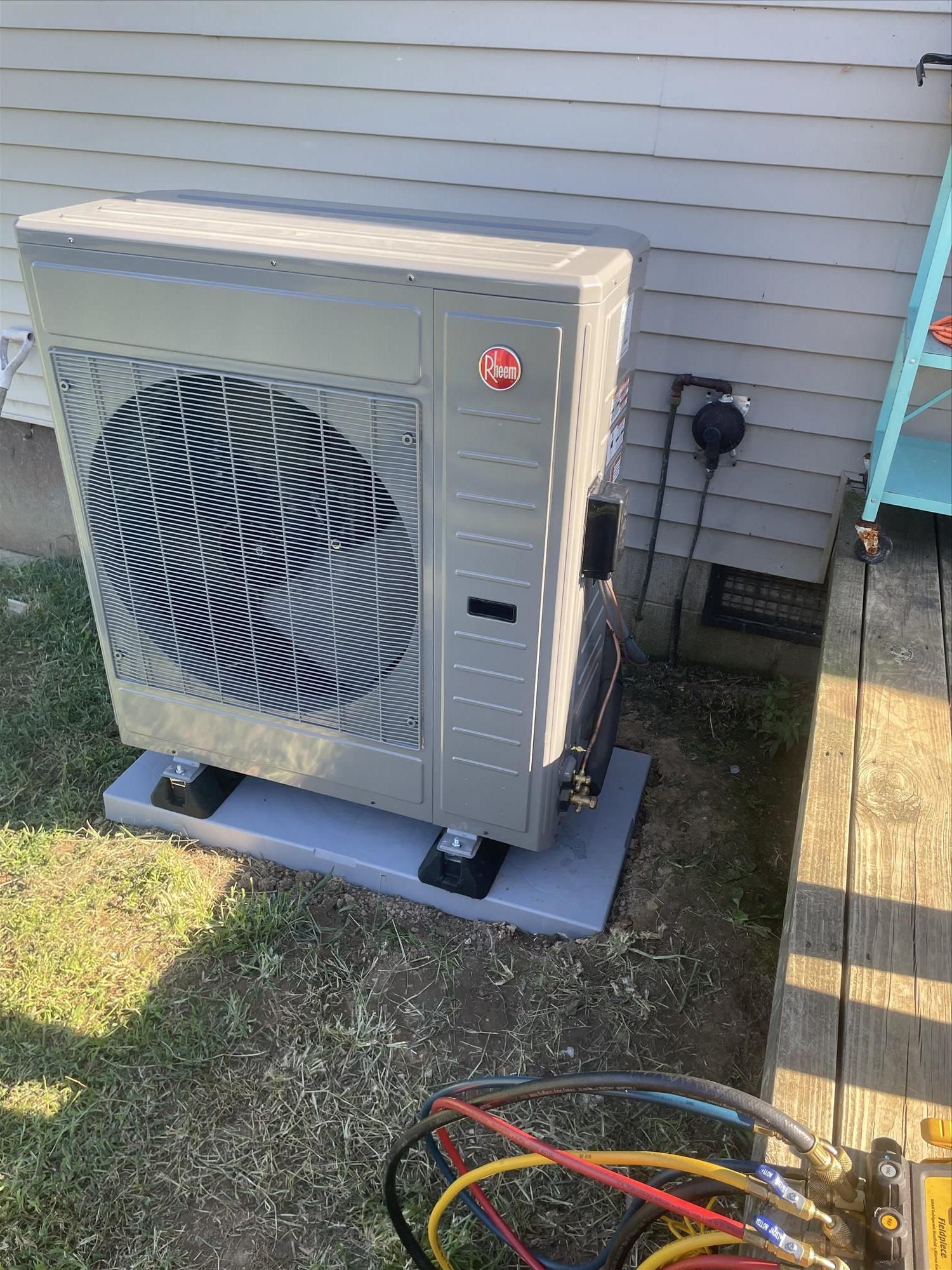 Rheem heat pump unit on gray platform next to a house. Hoses and gauges are in foreground.