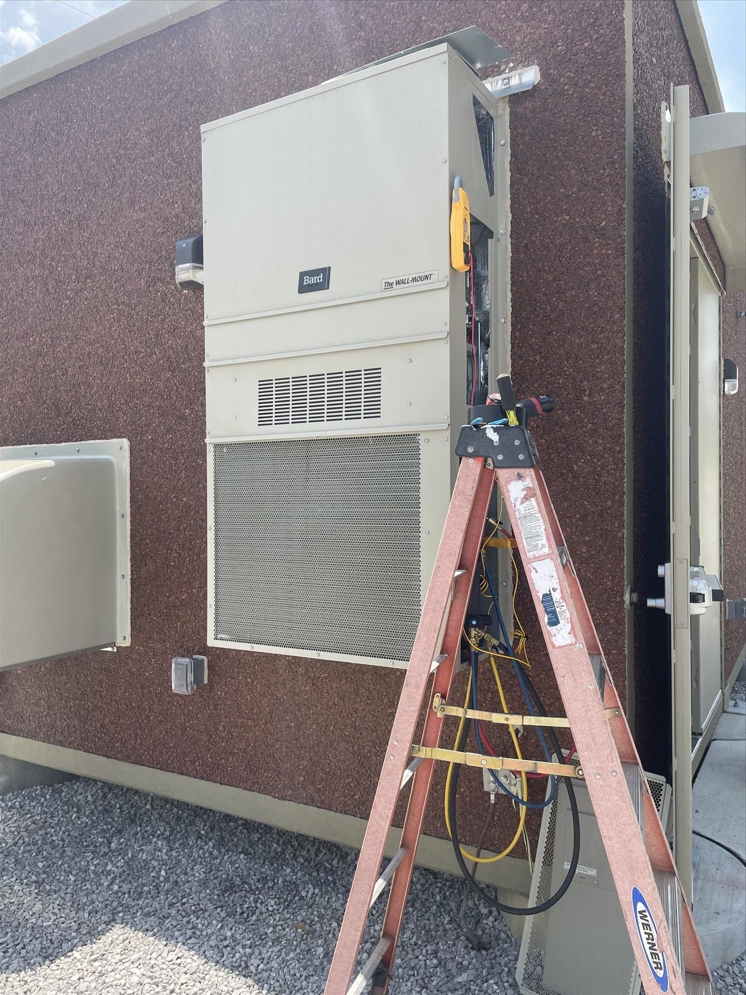 Air conditioning unit on brick building exterior, with ladder and open access panel for servicing.
