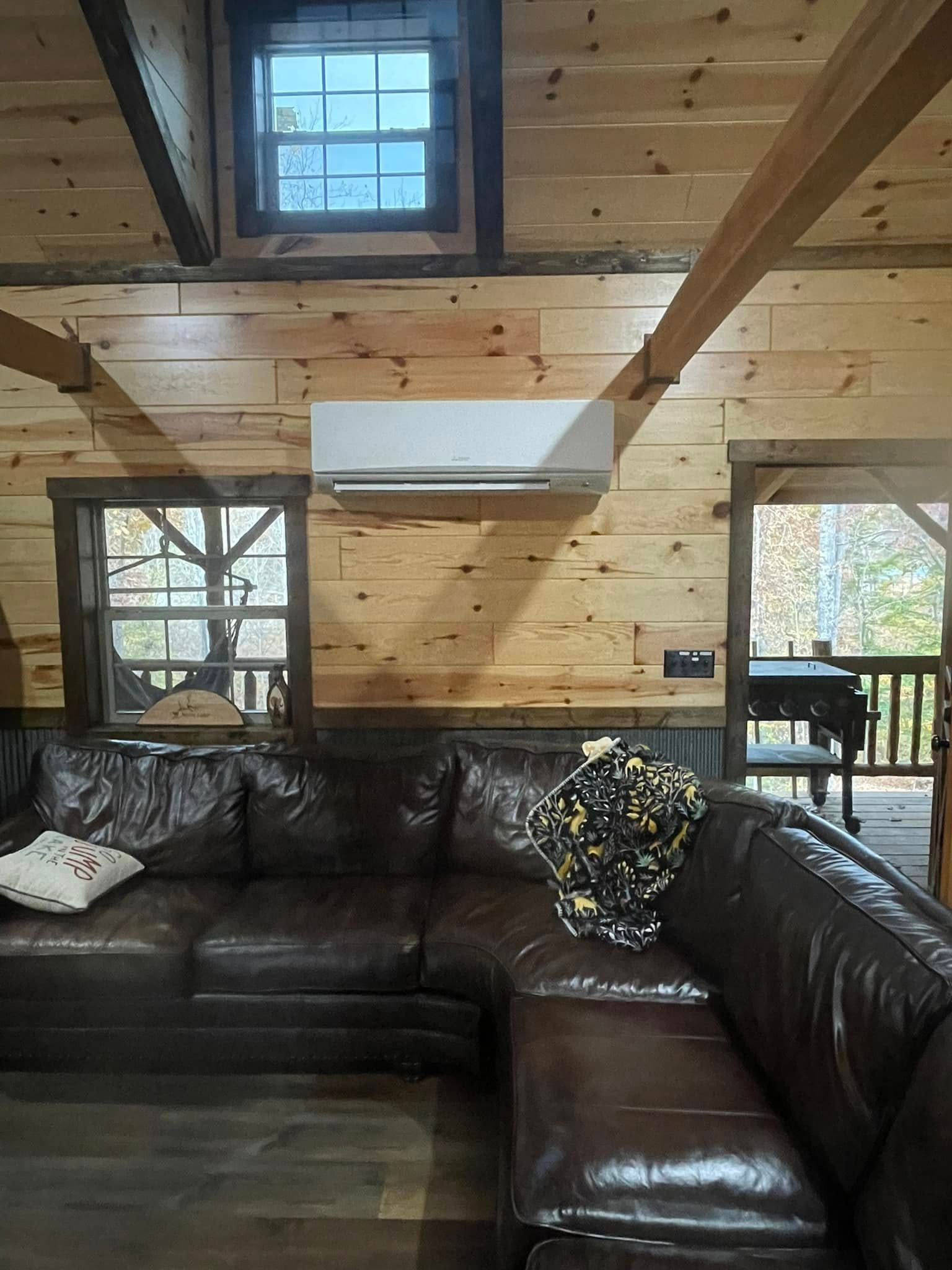 Cozy cabin interior: brown leather sectional, wood walls, small window, air conditioner.