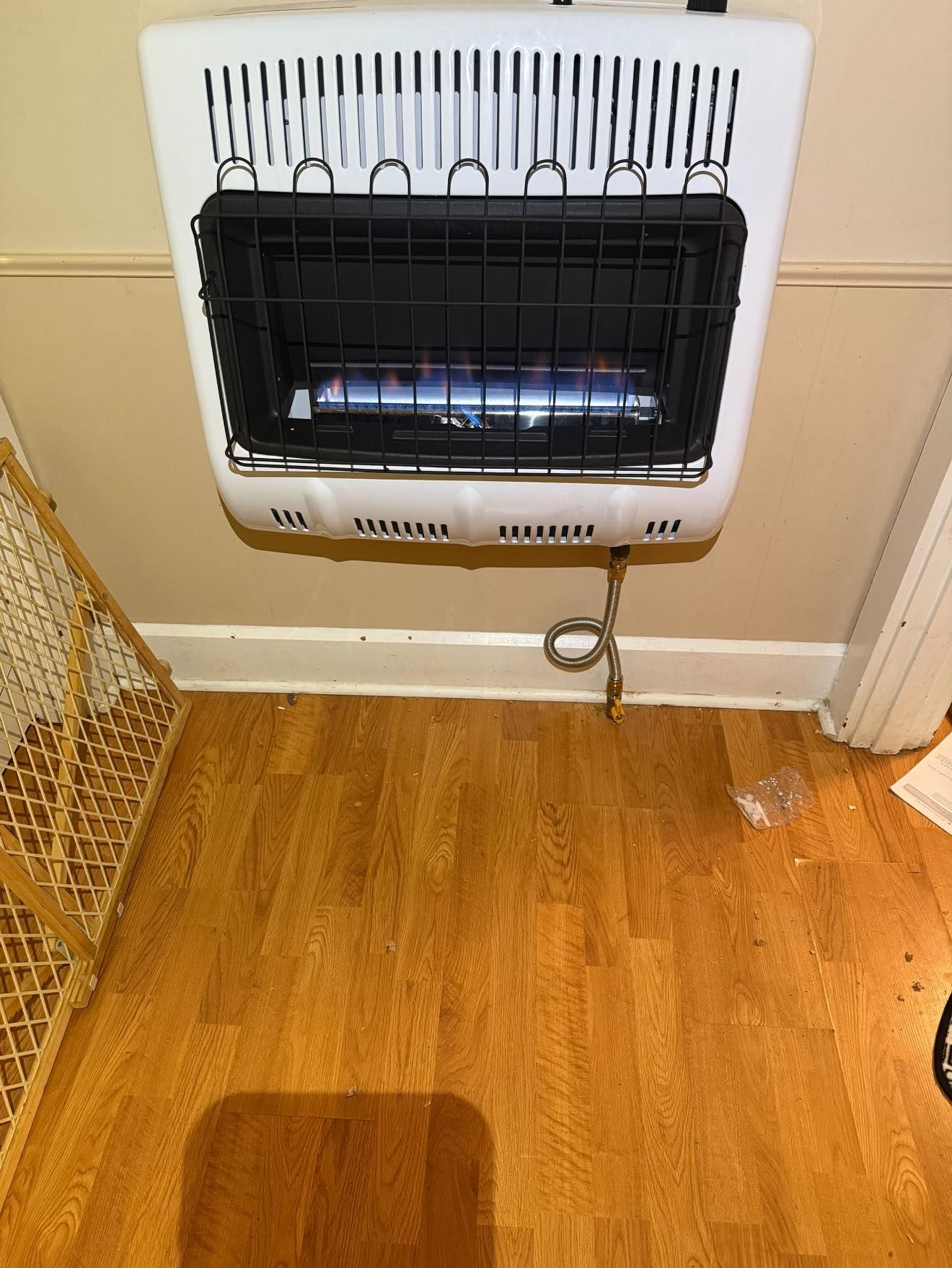 Wall-mounted gas heater with blue flames, attached to a beige wall above a hardwood floor.