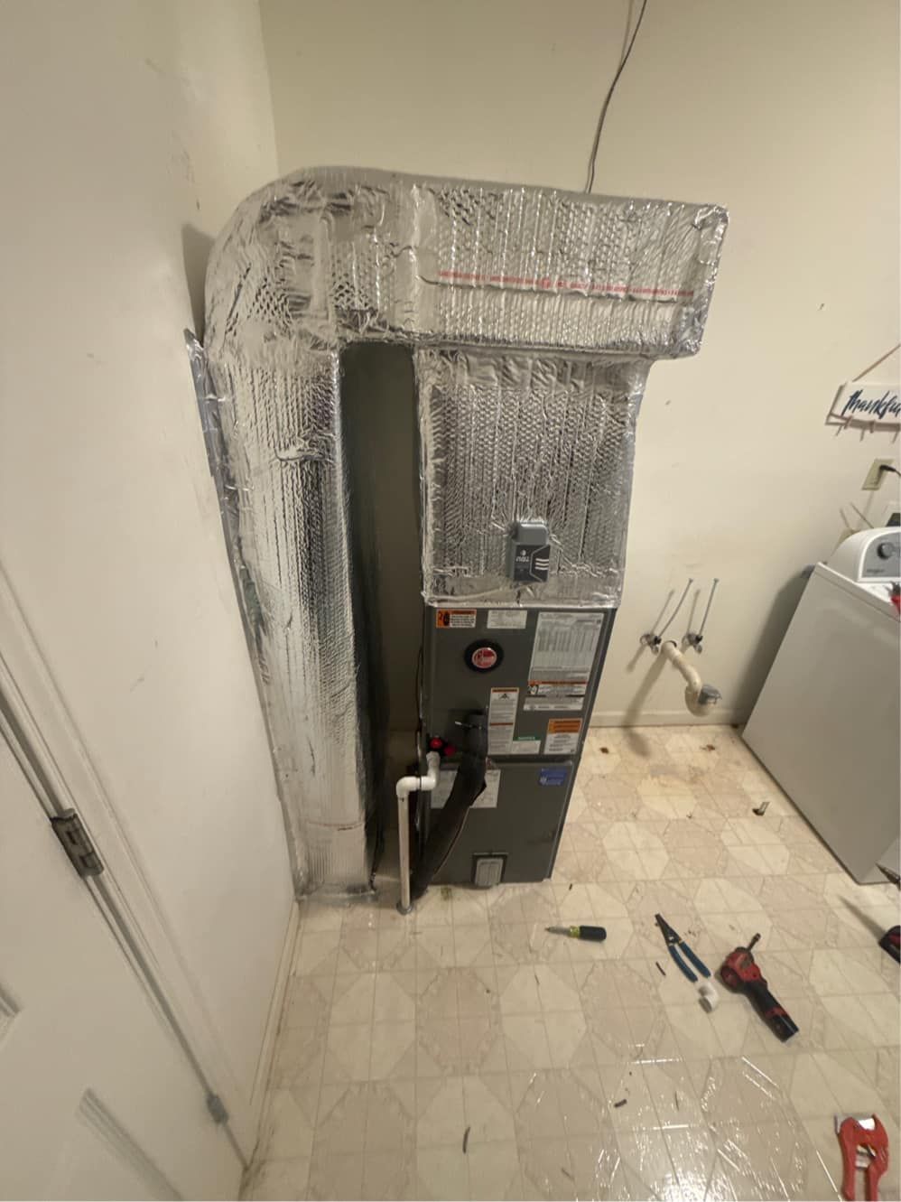 HVAC unit installation. Silver insulated ducts and unit against a white wall. Tools on floor.