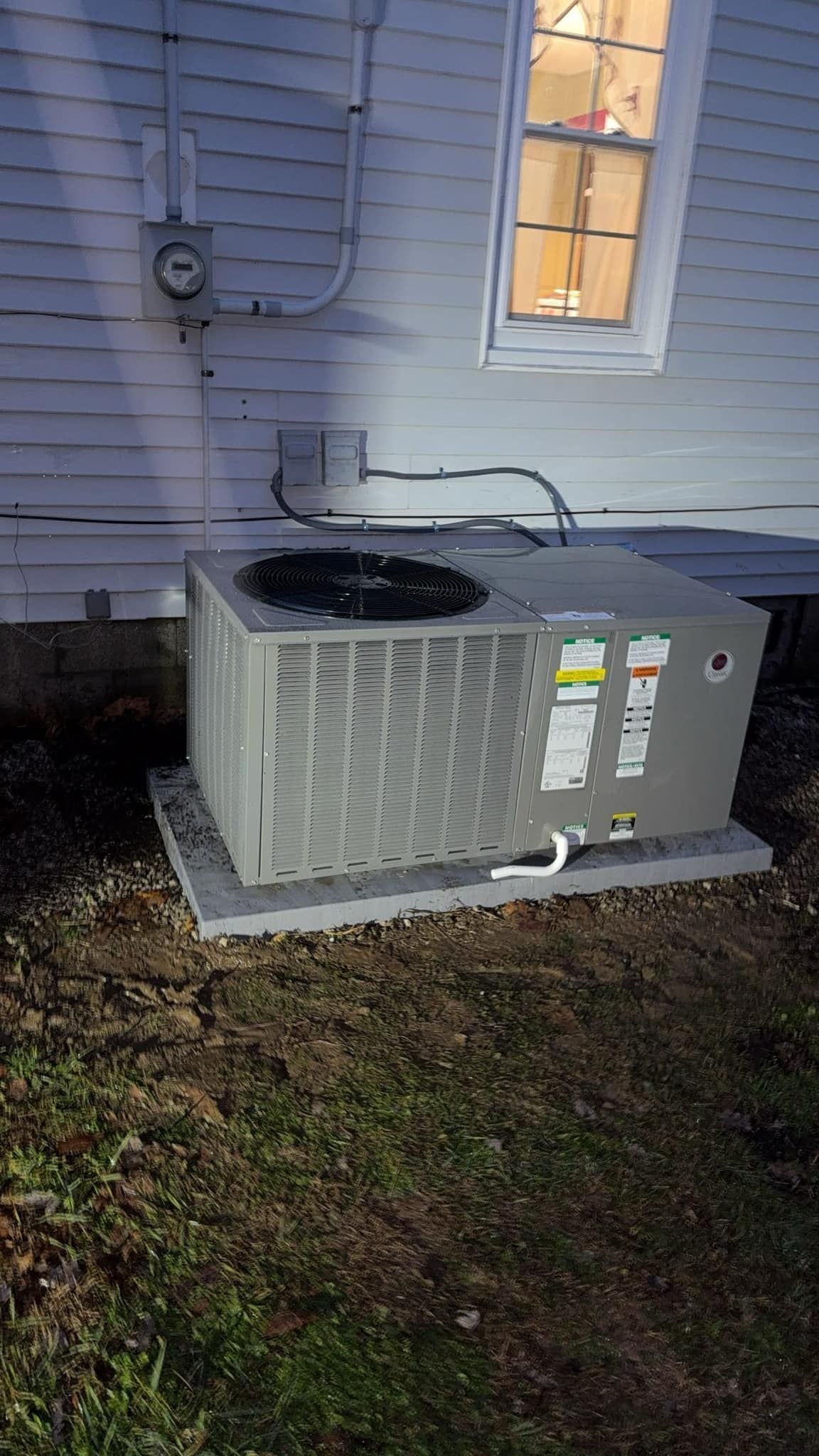 Air conditioner unit on a concrete pad next to a white house with a lit window.