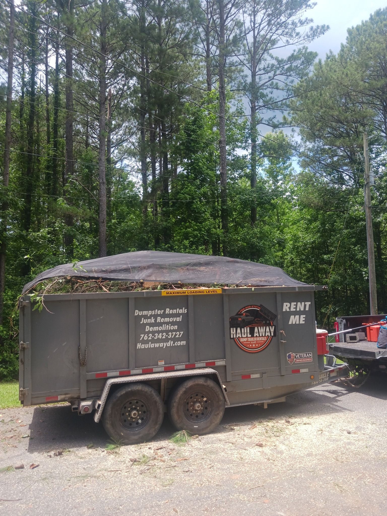 Gray trailer filled with green debris, parked near trees.