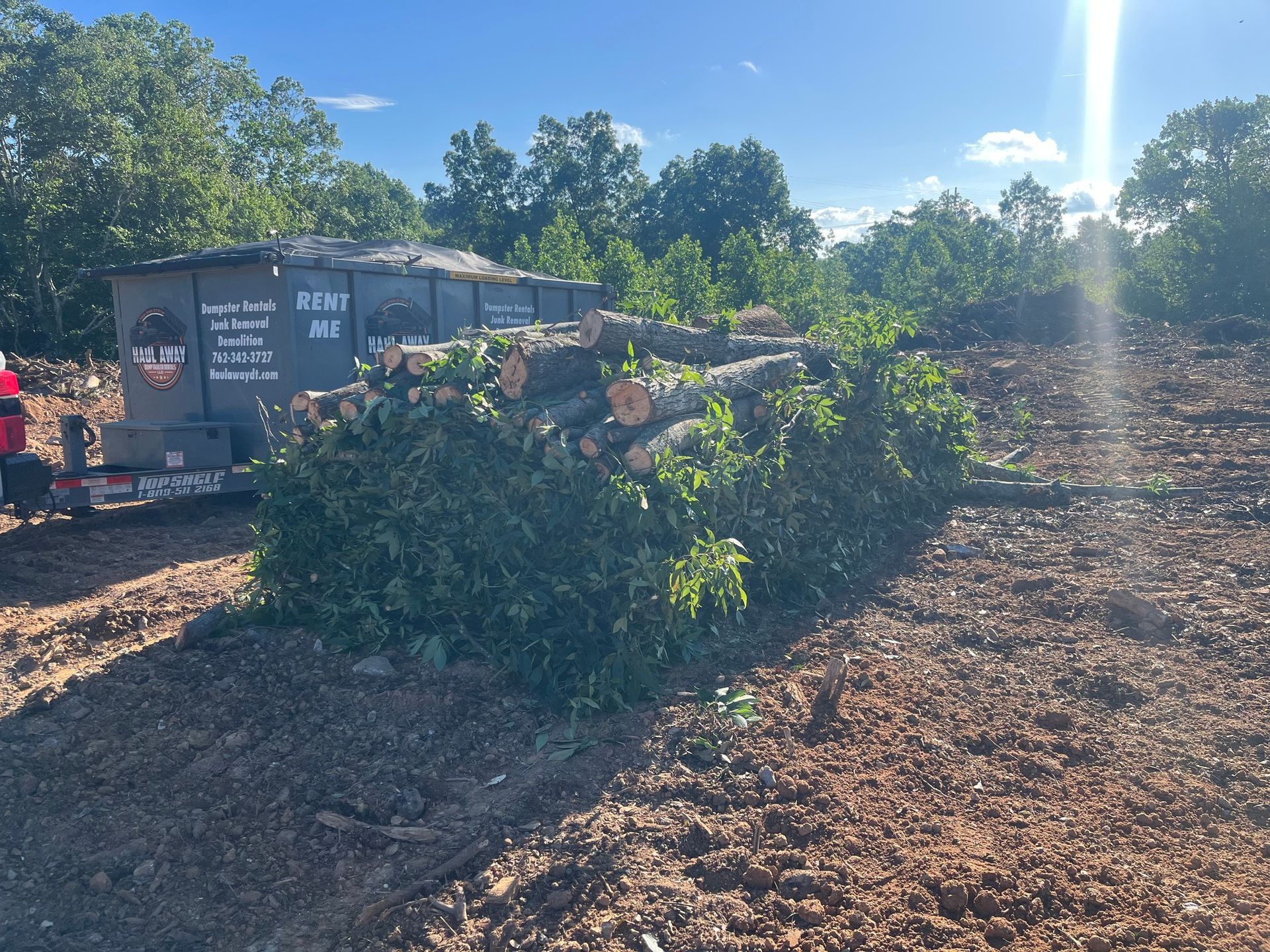 A pile of logs and debris next to a trailer in a cleared field with trees in the background under a sunny sky.