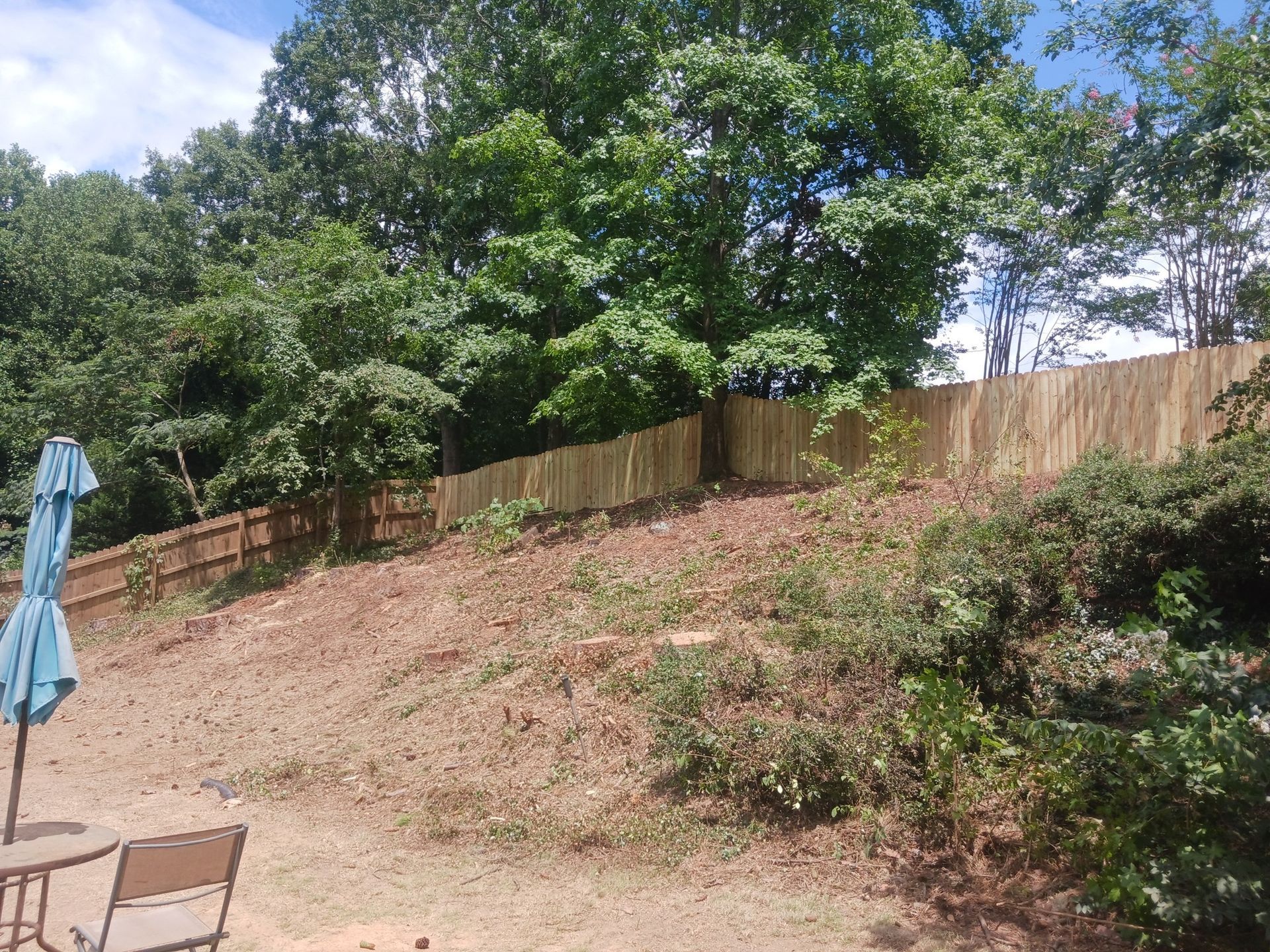 A wooden fence on a sloped yard with trees in the background under a blue sky.