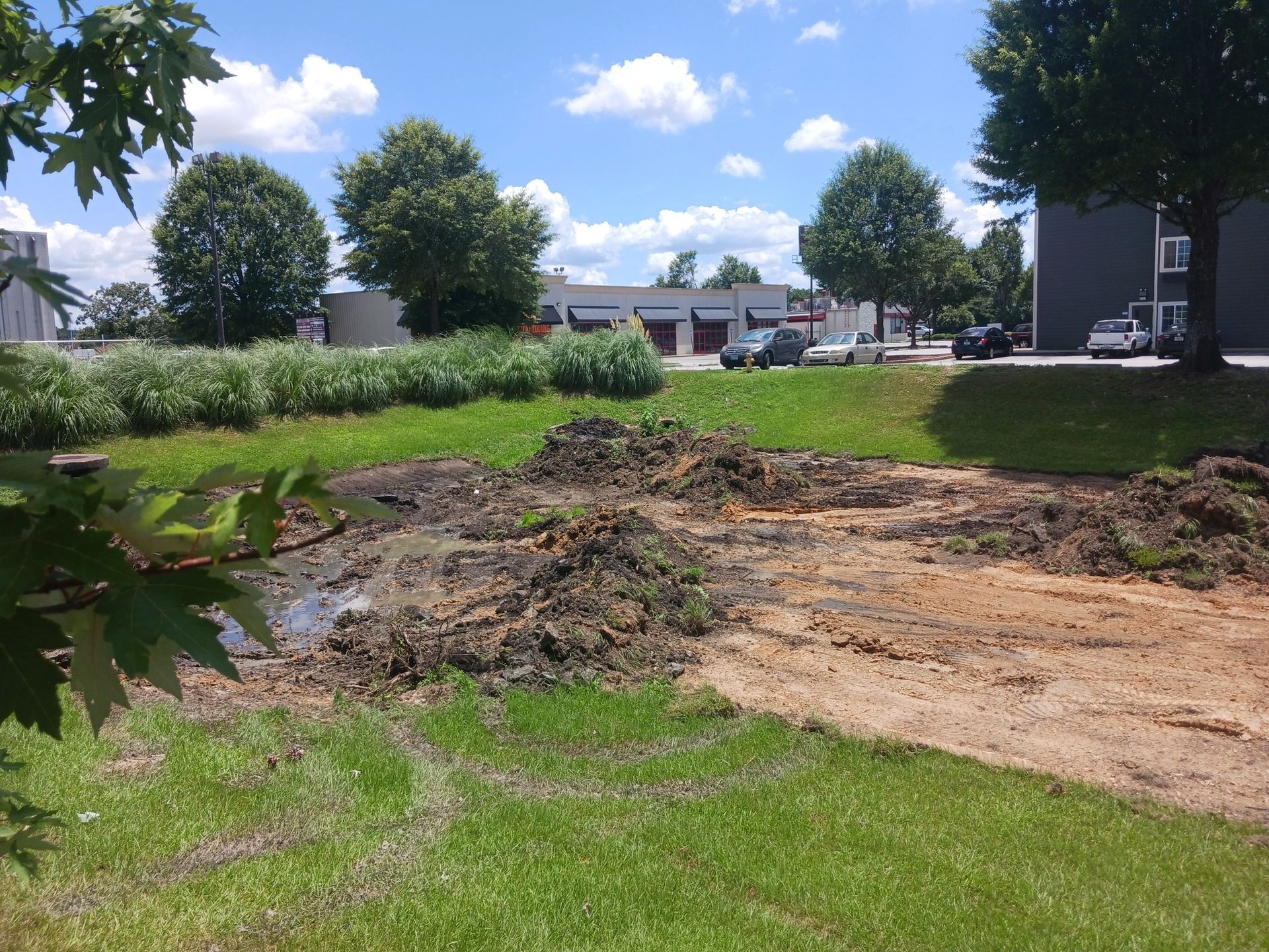 A muddy construction site in a grassy area with vehicles and buildings in the background under a blue sky.