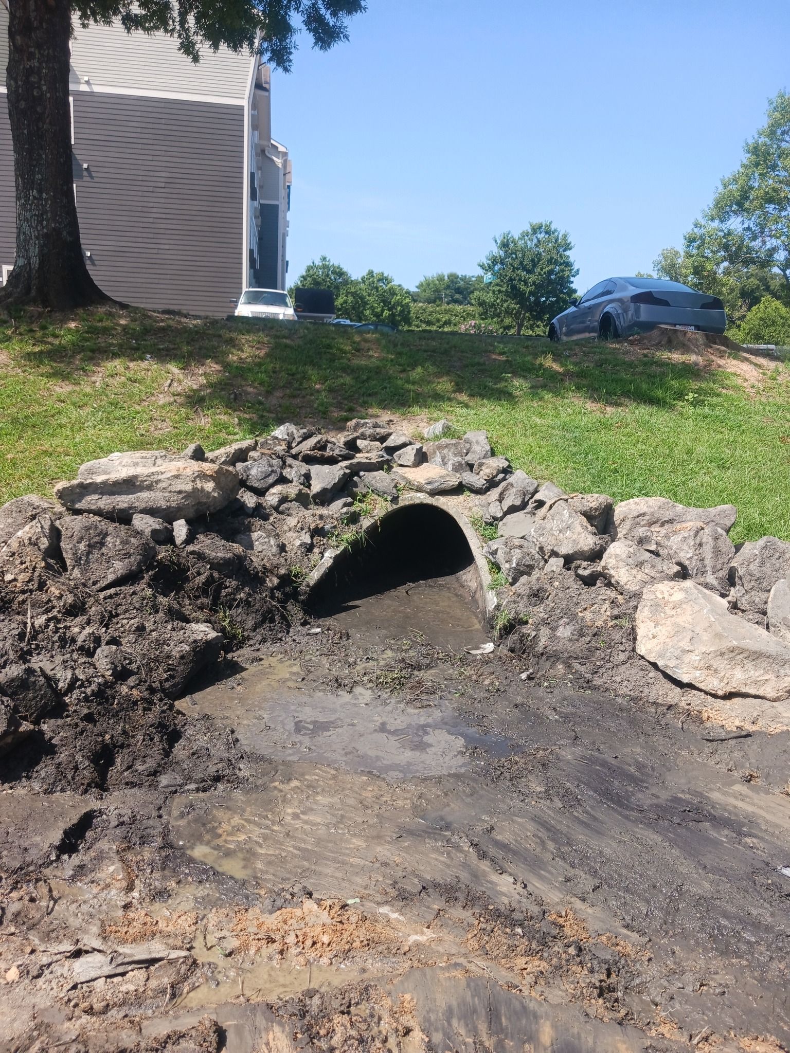 Stone arch over a dark drainage tunnel, set in dirt and surrounded by grass and a building.