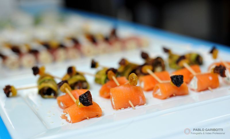 Aperitivos: rollitos de salmón con palillos, junto a otros alimentos enrollados en una bandeja blanca.