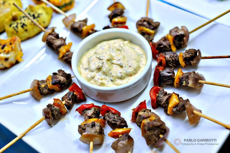 Carne y verduras en brochetas dispuestas alrededor de un pequeño recipiente con salsa en un plato blanco.