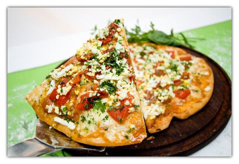 Porción de pizza con ingredientes, servida en un plato de madera, apoyada sobre una espátula de plata.