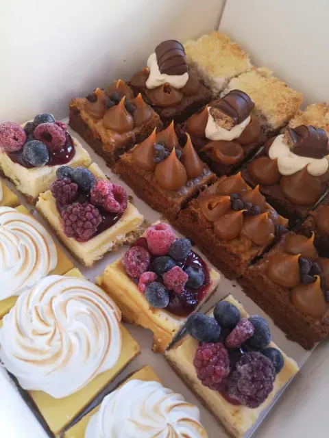 Caja de pasteles variados, que incluye cuadrados de tarta de queso con frutos rojos y merengue cubiertos con chocolate.