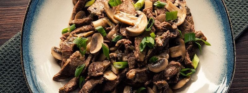 Salteado de carne de res y champiñones cubierto con cebollas verdes en un plato con borde azul.