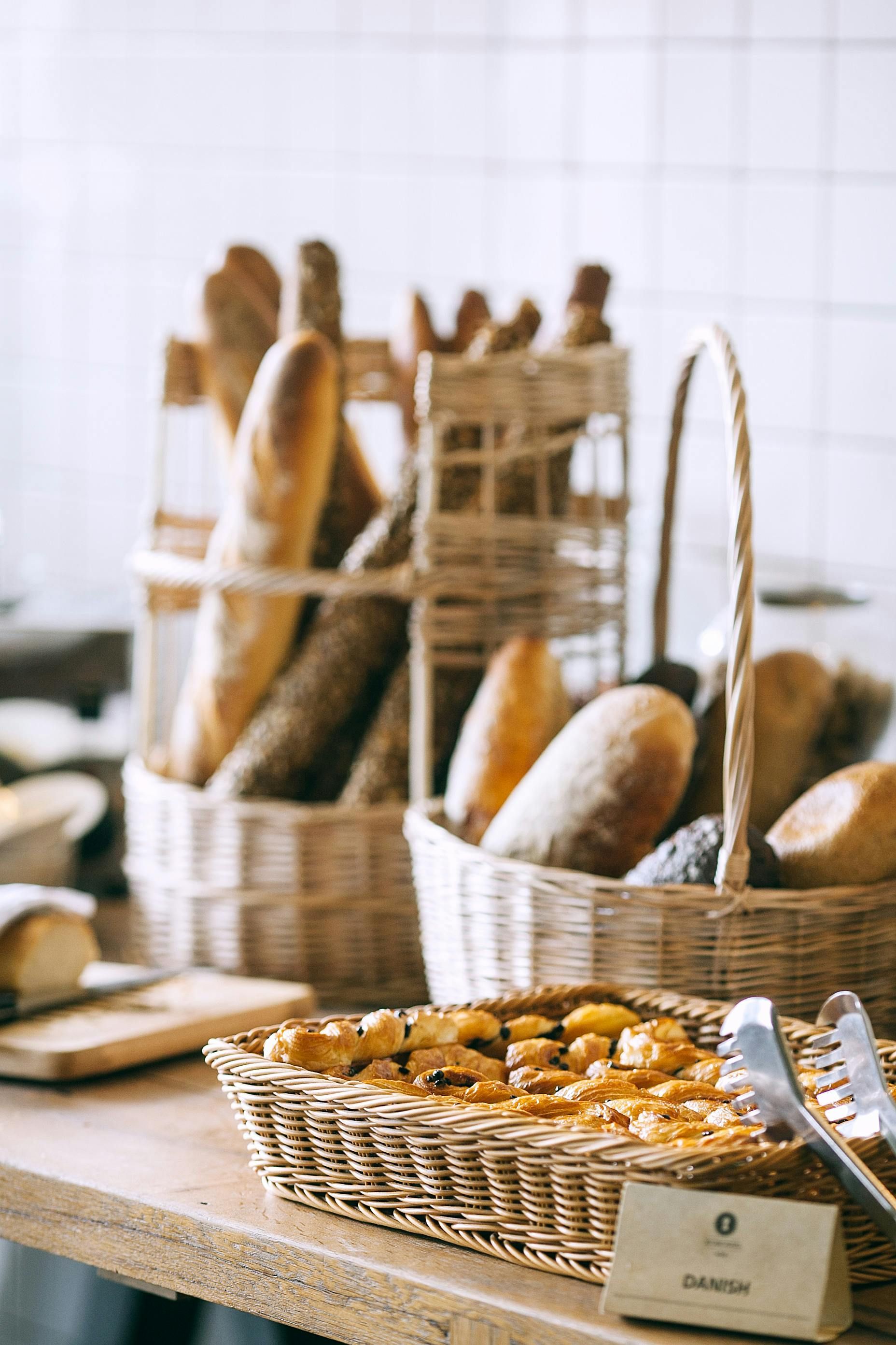 Cestas de panes y pasteles variados sobre un mostrador de madera, con pinzas, en un entorno luminoso.