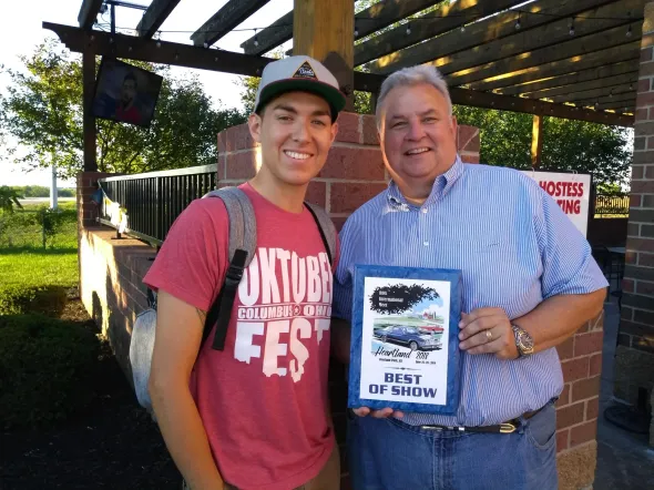 Two men smiling, holding an award outside. One wears a red shirt that says