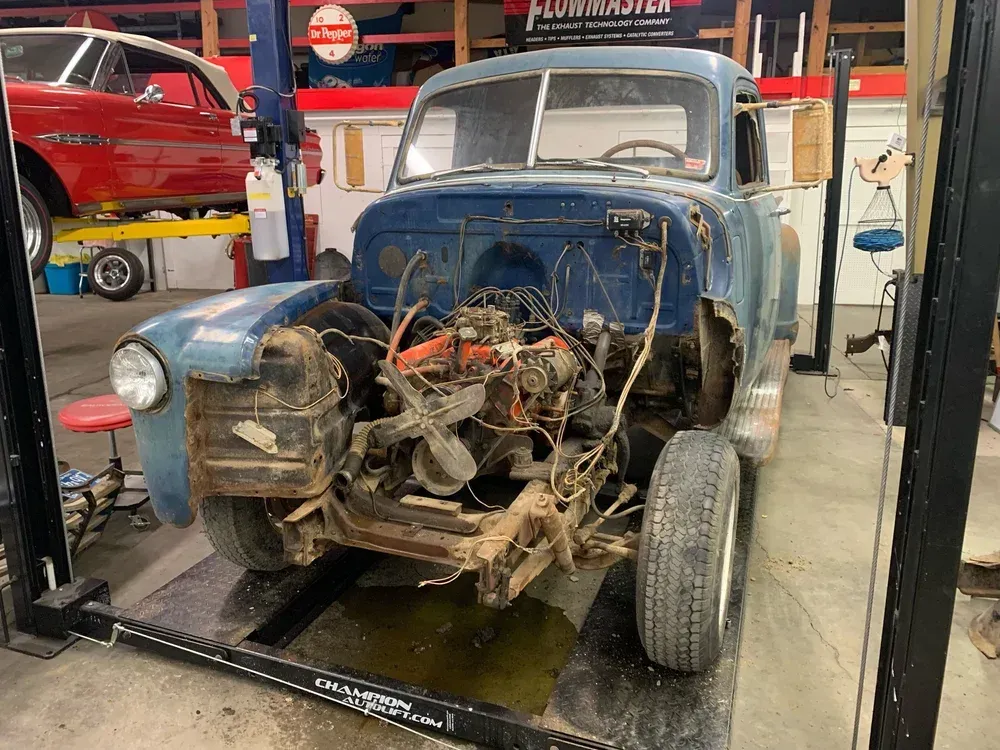 Blue vintage truck undergoing restoration in a garage. Engine exposed, other car in background on a lift.