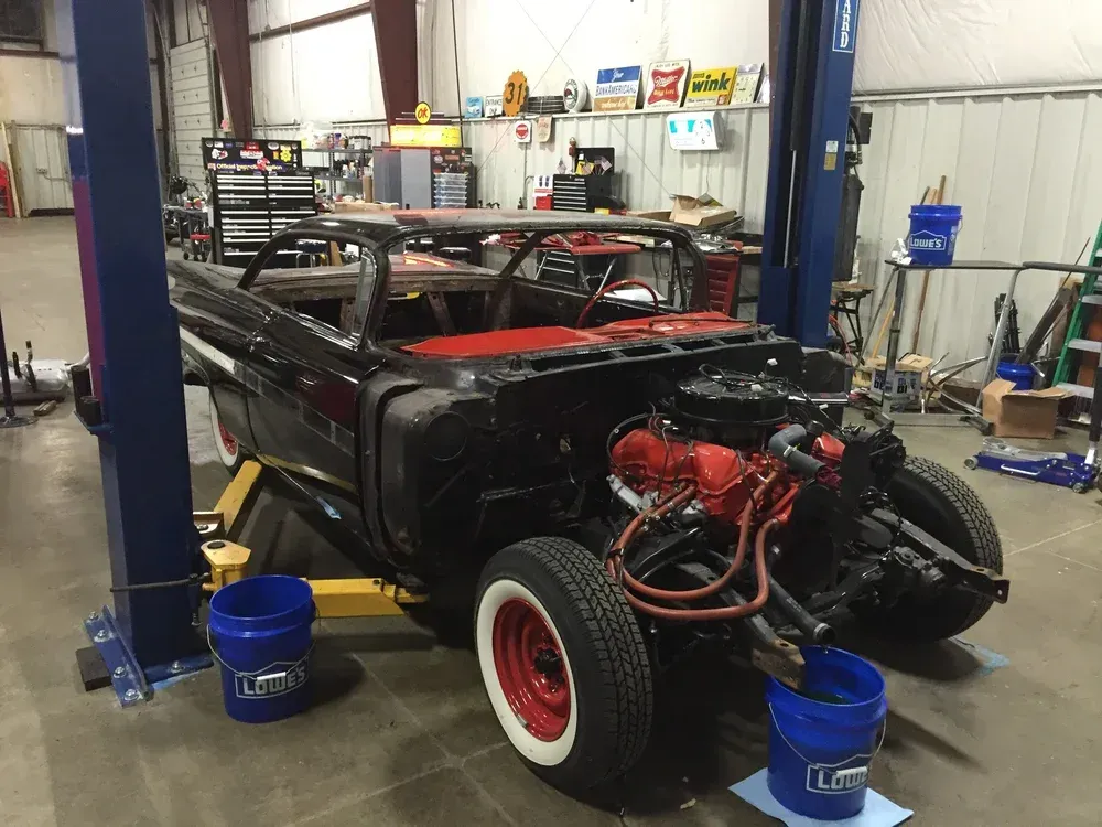 Black classic car in a garage, engine exposed, up on a lift.