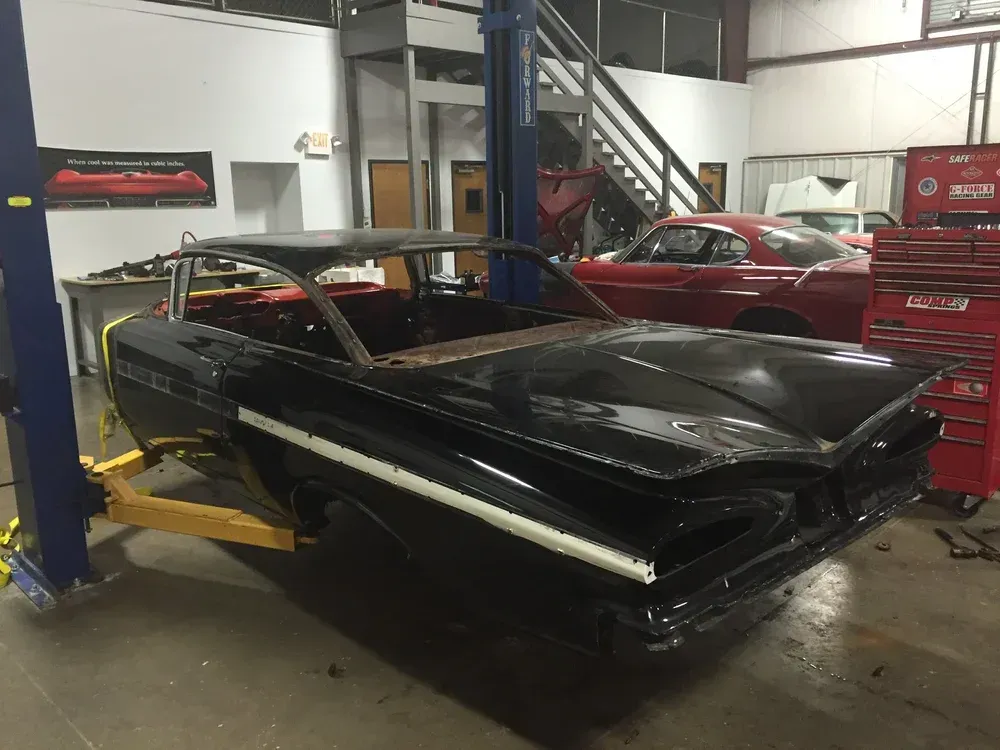 Black classic car on a lift in a garage. Another red car is in the background. Tools and stairs are visible.