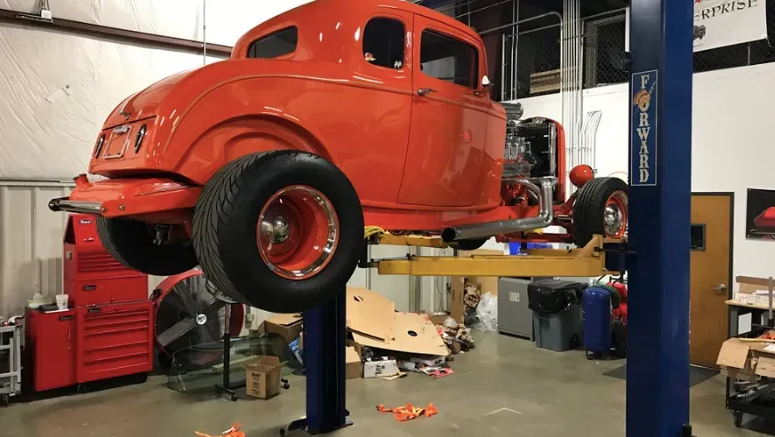 Orange hot rod coupe on a car lift in a garage, with large rear tires and a visible engine.