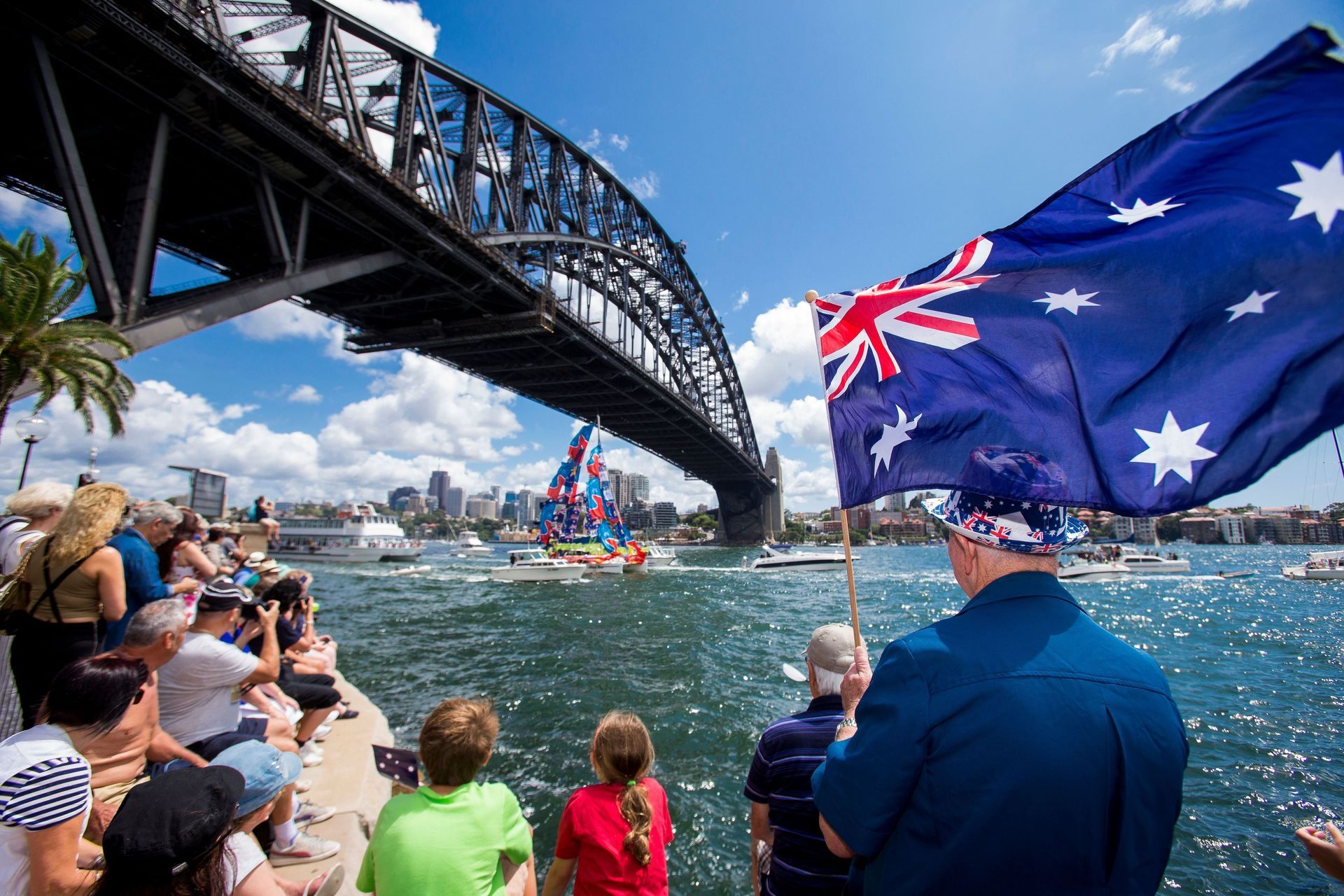 Crowds celebrate Australia Day under the Sydney Harbour Bridge, watching boats decorated with flags 