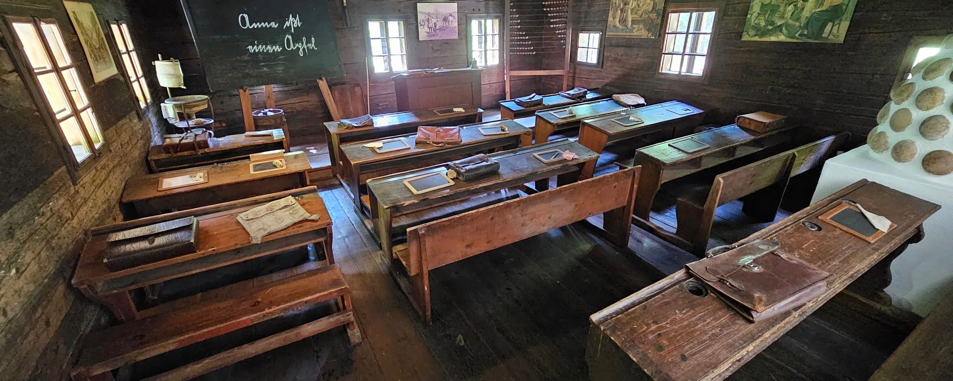 Interior of a restored 19th-century Australian bush school classroom featuring timber walls, wooden desks and benches, old slates and leather satchels on the desks, and a chalkboard at the front. Natural light filters through small windows, giving a glimpse into early rural education.